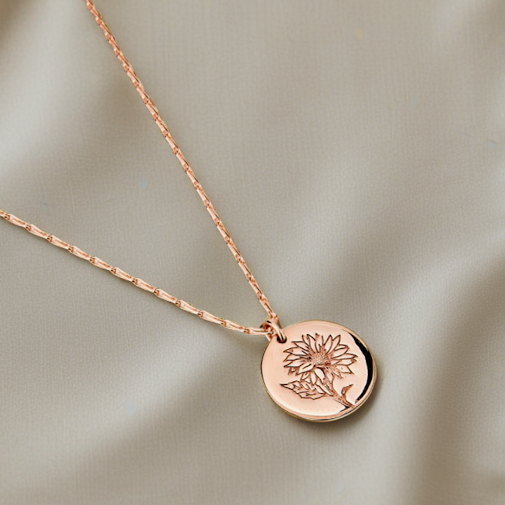 Sunflower Medallion necklace in rose gold featuring a round engraved sunflower pendant on a delicate chain, displayed against a soft neutral fabric background.