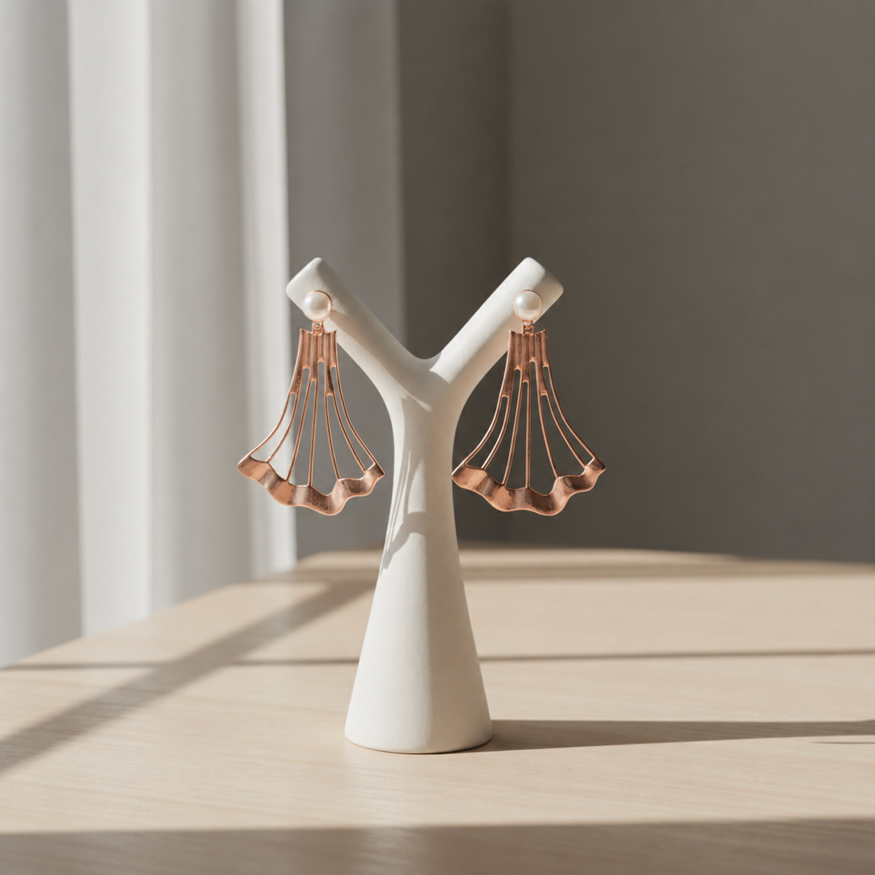 Handcrafted Blush Tide shoulder duster earrings in rose gold plated brass displayed on a minimal white sculptural stand, featuring a fan-shaped architectural design topped with real freshwater pearls.