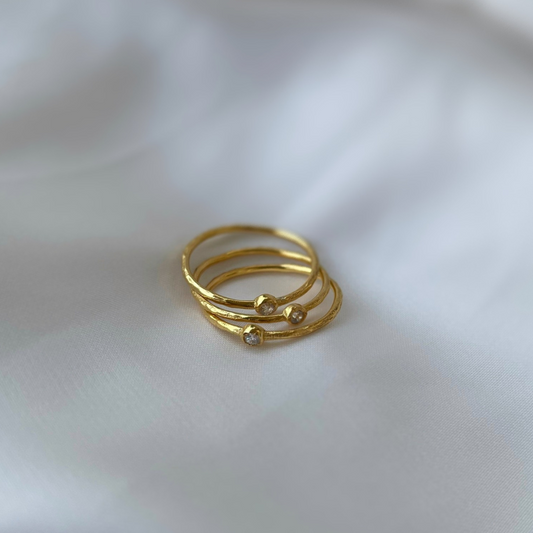 Three gold-plated stackable rings with a rustic hammered texture and stone accents, artistically arranged on a soft white fabric surface.
