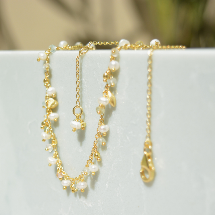 The Natural Grace pearl and granite charm necklace draped over a minimal white stone block, showcasing the playful movement of the drop charms and the warm golden finish.