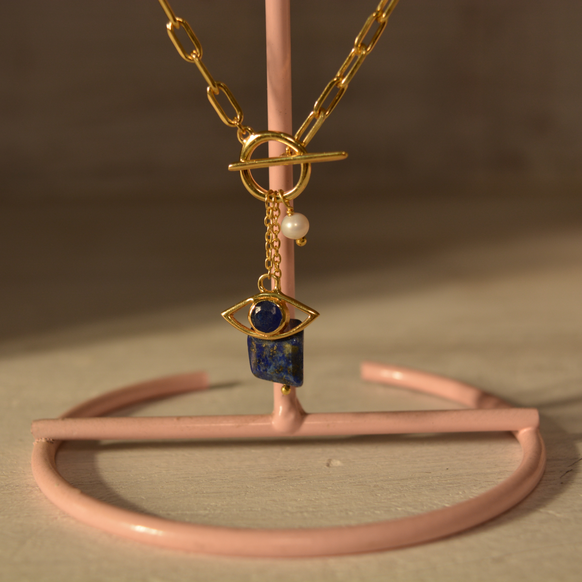 Detail shot of the Meaningful Charm Necklace hanging on a pink stand, highlighting the marquise-shaped lapis lazuli, evil eye protection symbol, and the textured paperclip chain.