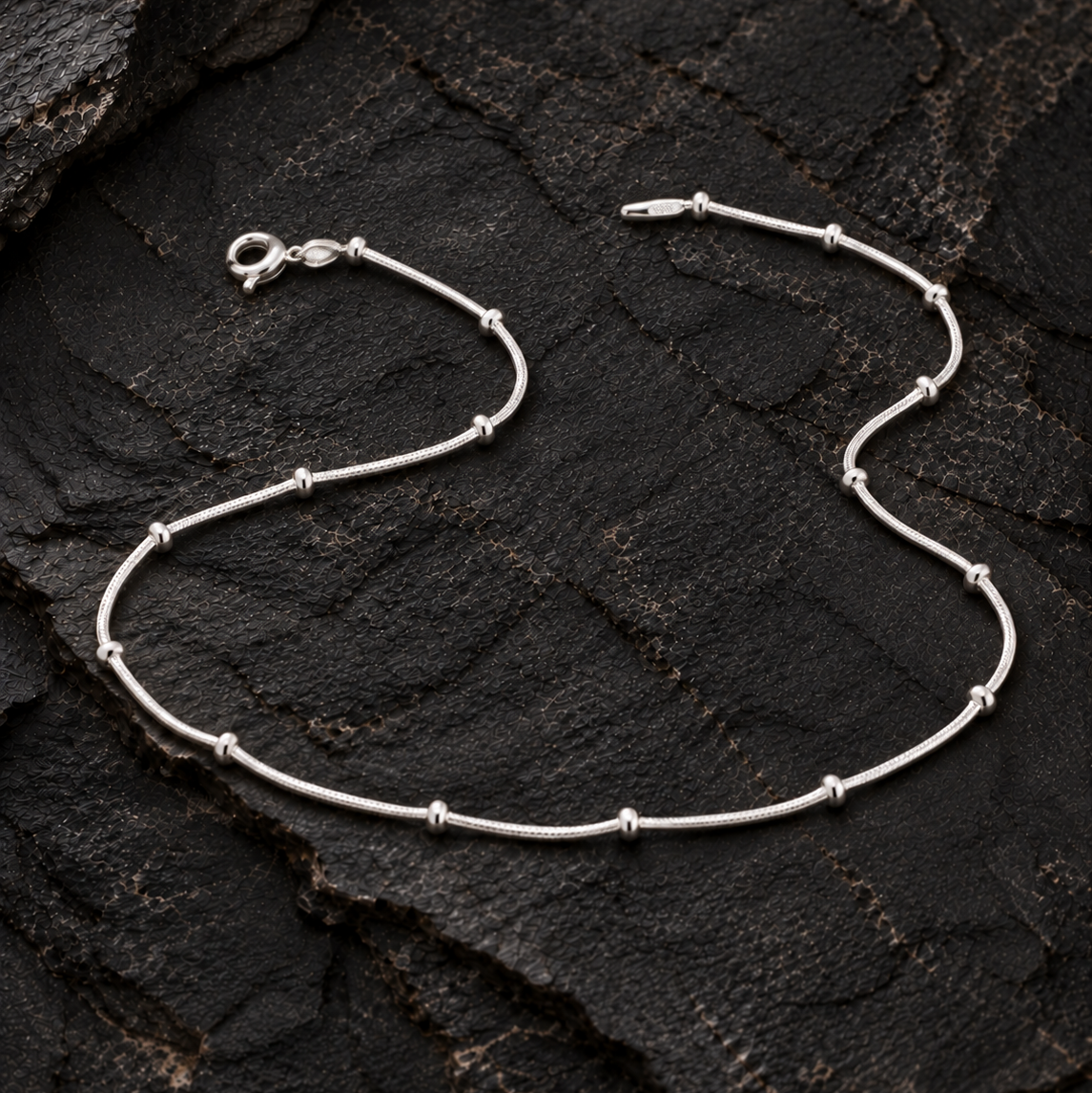 Silver anklet with spaced beads on a dark textured rock surface