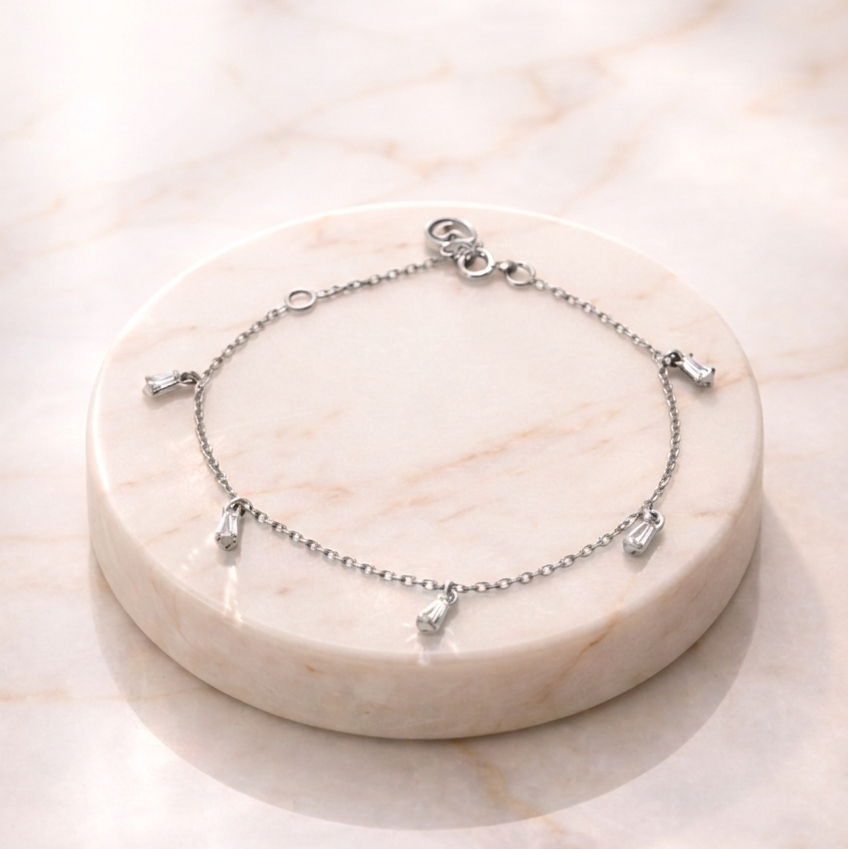 A delicate Floating Stone Bracelet featuring five small, sparkling baguette-cut stones hanging as charms from a fine cable chain, displayed on a circular white marble pedestal in soft, natural light.
