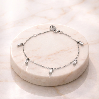 A delicate Floating Stone Bracelet featuring five small, sparkling baguette-cut stones hanging as charms from a fine cable chain, displayed on a circular white marble pedestal in soft, natural light.