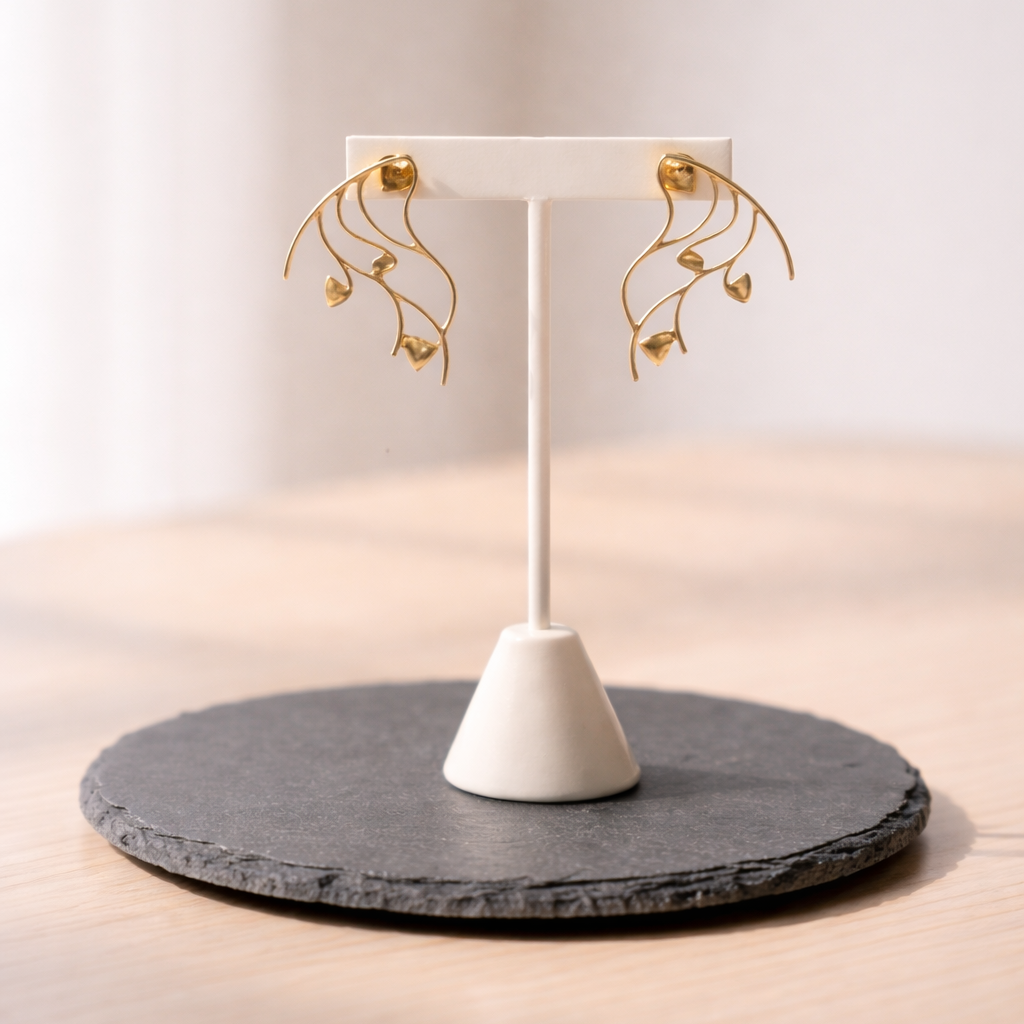 A pair of Delicate Golden Entangled Earrings featuring an intricate, wavy vine-like gold design with small leaf accents, displayed on a white T-bar jewelry stand atop a dark gray circular slate base in a soft, sunlit indoor setting.