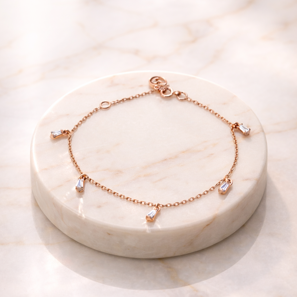 A delicate rose gold Floating Stone Bracelet featuring five small, sparkling baguette-cut stones hanging as charms from a fine cable chain, displayed on a circular white marble pedestal in soft, natural light.