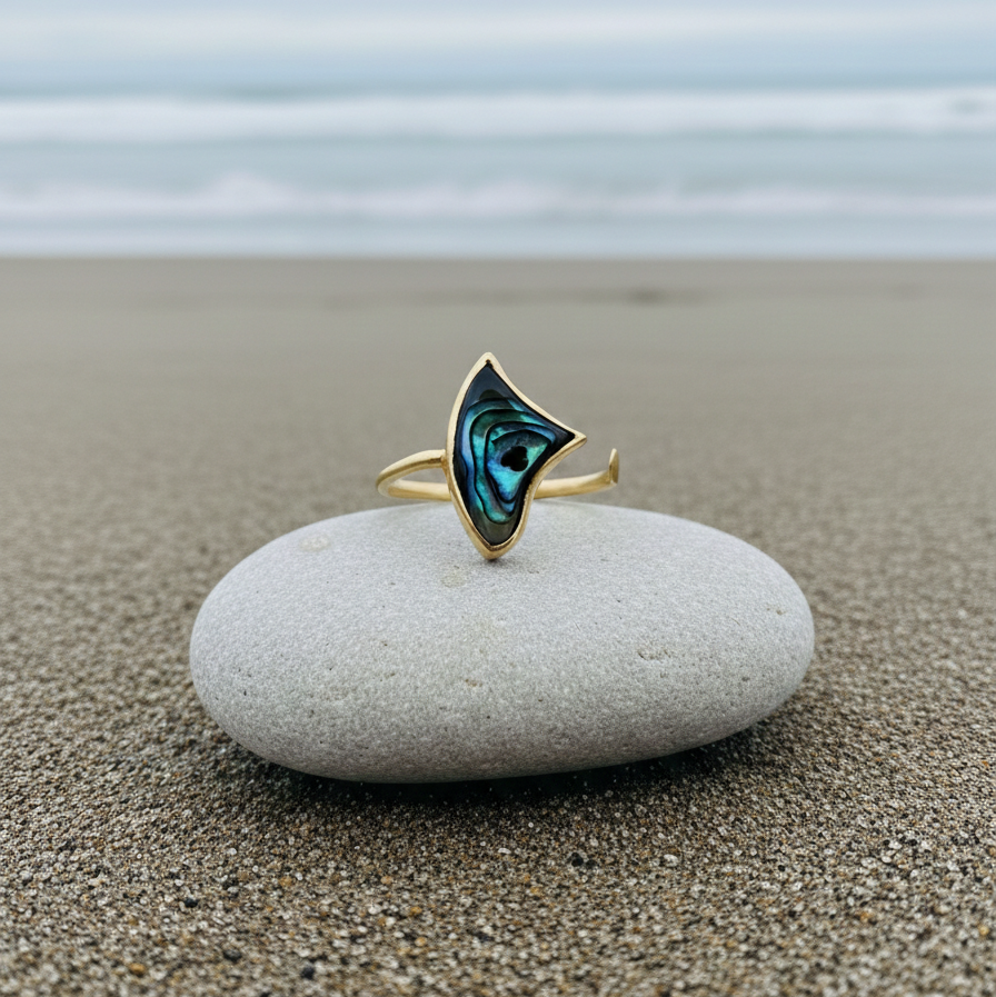 Adjustable open gold knuckle ring with luminous blue-green inlay detail, photographed on a stone with ocean in the background.
