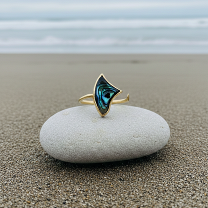 Adjustable open gold knuckle ring with luminous blue-green inlay detail, photographed on a stone with ocean in the background.