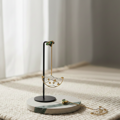 Long gold shoulder duster earring with delicate wave-like design and green accent displayed on a minimalist stand.