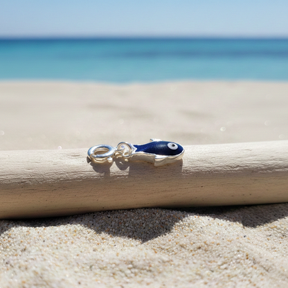 Hand-enamelled evil eye fish charm crafted in 925 silver resting on a wooden stick on sandy beach with blue ocean in the background.