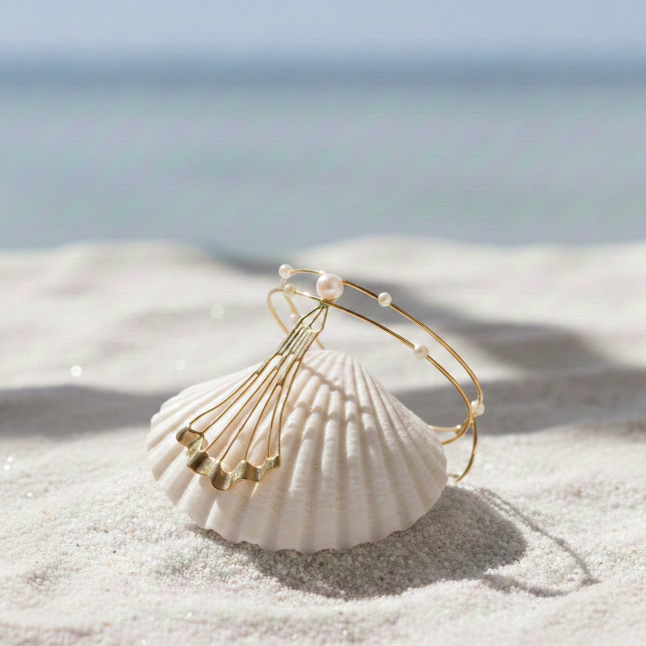 Modern gold cuff bracelet with flowing fan-shaped detail and pearls placed on a seashell in natural coastal lighting.