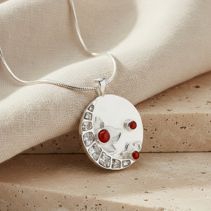 Full Moon Pendant in silver-plated finish featuring red stone accents and CZ details, styled on a chain against a neutral fabric and stone backdrop.