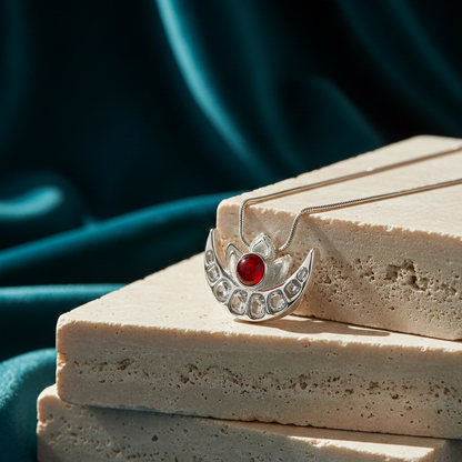 Silver half moon pendant necklace featuring a red gemstone centerpiece and crescent design displayed on stacked stone blocks against a teal background.