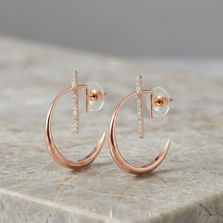 A pair of La Luna Rose Gold Earrings featuring a polished crescent moon hoop design with a diamond-studded vertical bar, displayed on a light-colored marble surface.