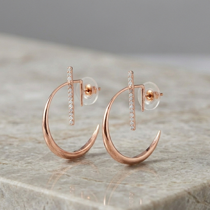 A pair of La Luna Rose Gold Earrings featuring a polished crescent moon hoop design with a diamond-studded vertical bar, displayed on a light-colored marble surface.