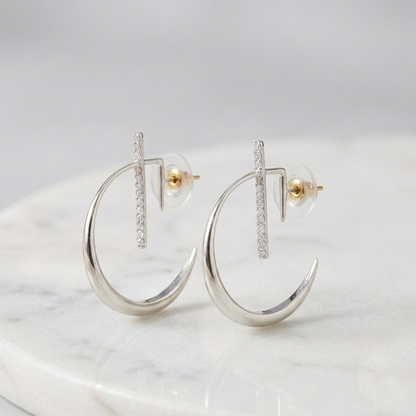 A pair of La Luna Platinum Earrings featuring a sleek, high-polished crescent moon hoop design intersected by a vertical bar set with shimmering pavé diamonds, displayed on a white marble surface.