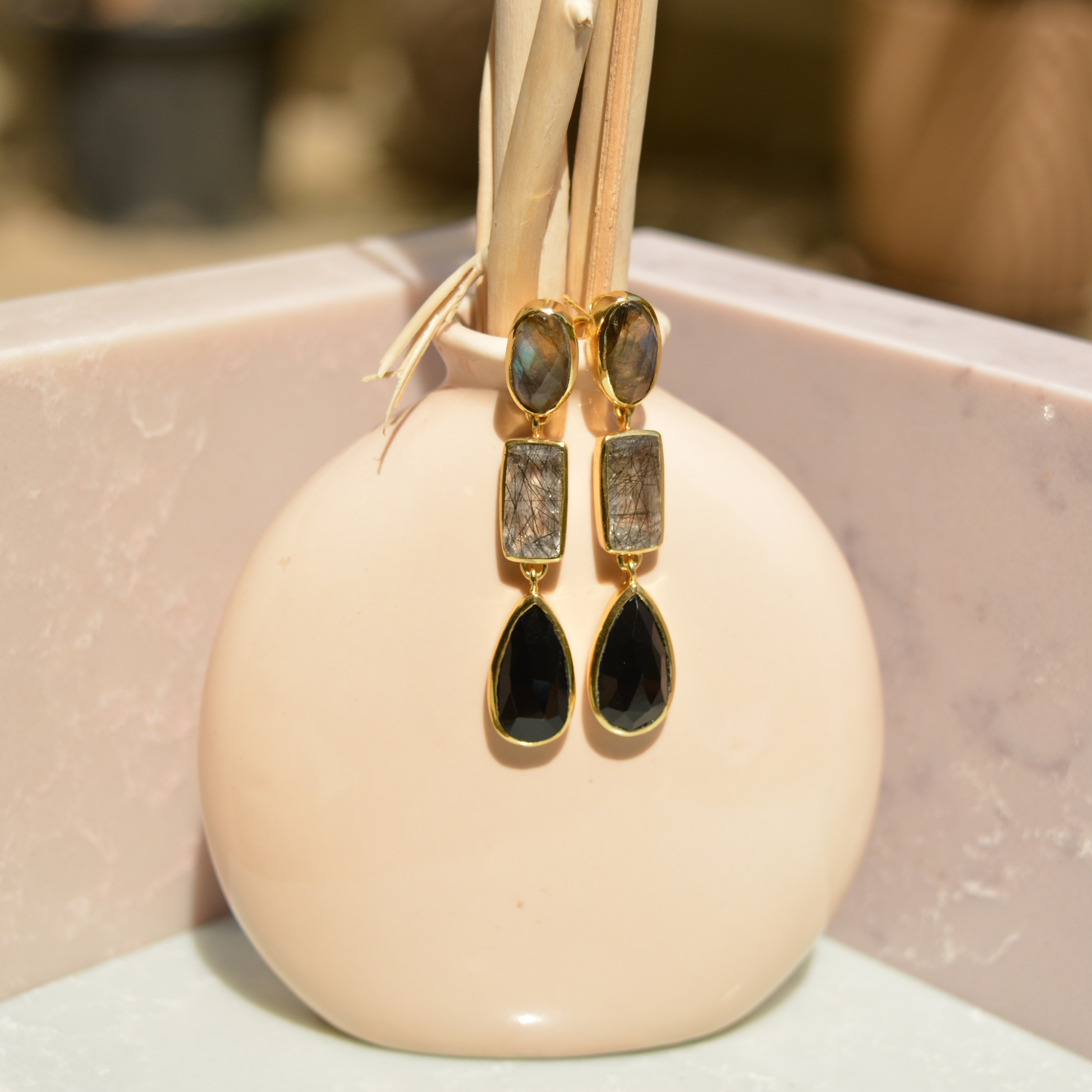 Lifestyle view of Midnight Elegance long drop earrings featuring Labradorite, Black Rutile, and Black Onyx set in 18kt gold-plated silver on a minimal vase.