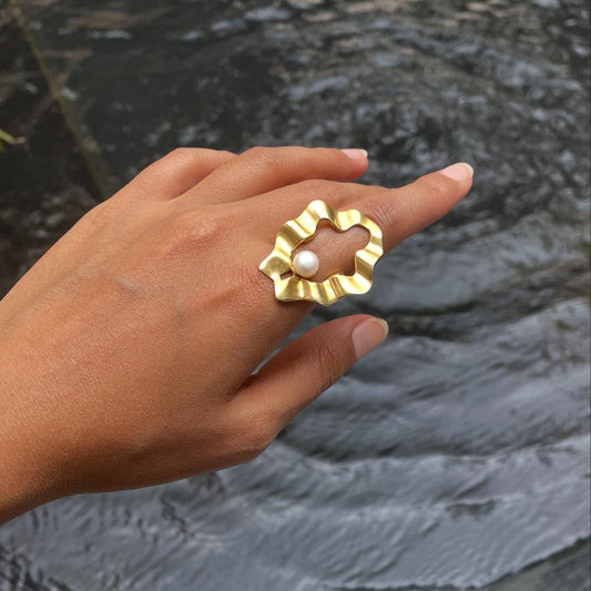 An outdoor lifestyle shot of a hand wearing an organic, wavy gold ring with a central pearl against a water background.