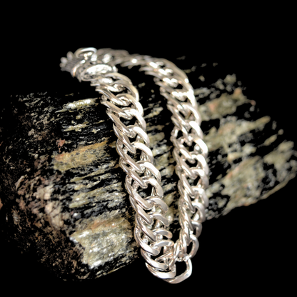 Close-up of a heavy, polished silver-tone chain bracelet draped over a textured stone, highlighting the intricate double-link weave and masculine style.