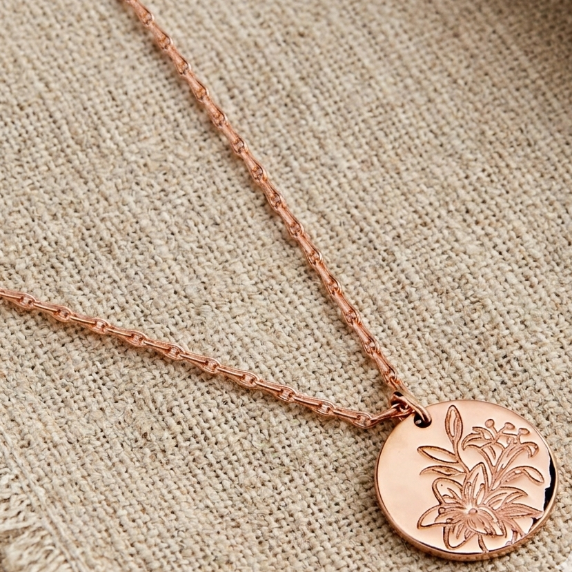 Lily Medallion Necklace in rose gold featuring a round engraved floral pendant on a delicate chain, displayed on textured linen fabric.