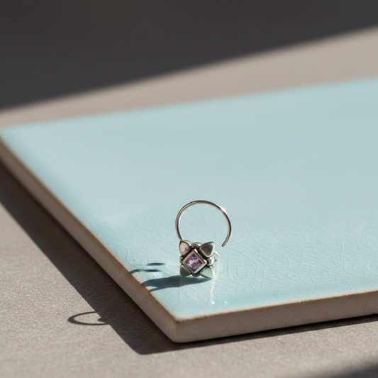 Silver nose pin with floral design and square-cut purple gemstone displayed on a soft blue tile in natural light.