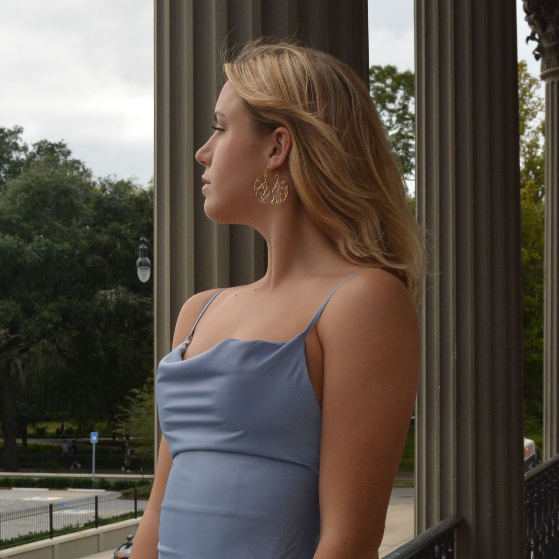 Profile view of a blonde model wearing a light blue dress and two-tone circular drop earrings, standing on a balcony with classical columns in the background.