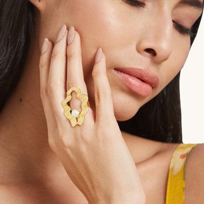 A portrait of a model touching her face, highlighting a sculptural gold ring with a delicate pearl centerpiece on her finger.