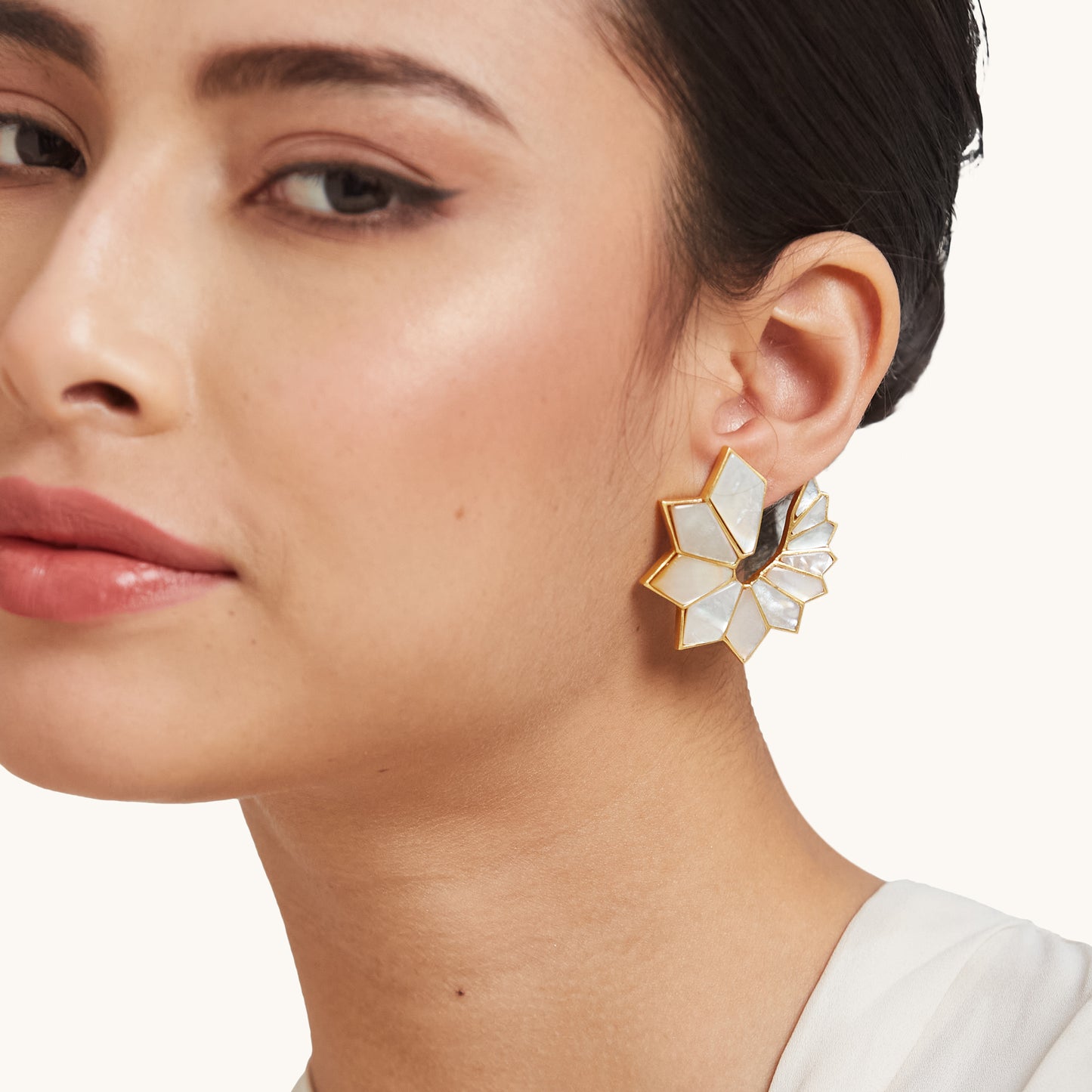 A model showcasing a bold, sunburst-style geometric earring with iridescent white panels and a gold border against a clean studio background.