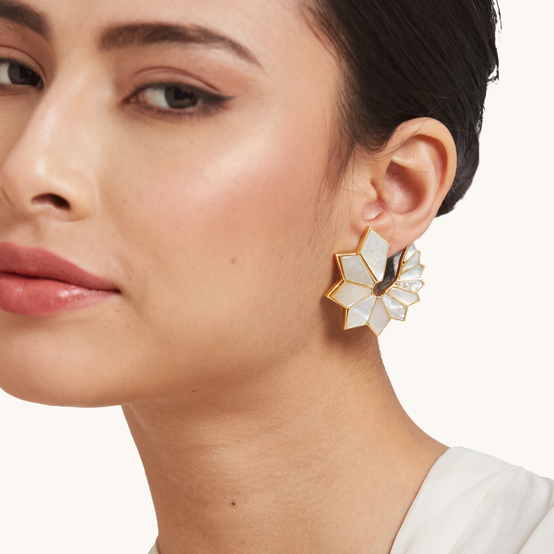 A model showcasing a bold, sunburst-style geometric earring with iridescent white panels and a gold border against a clean studio background.