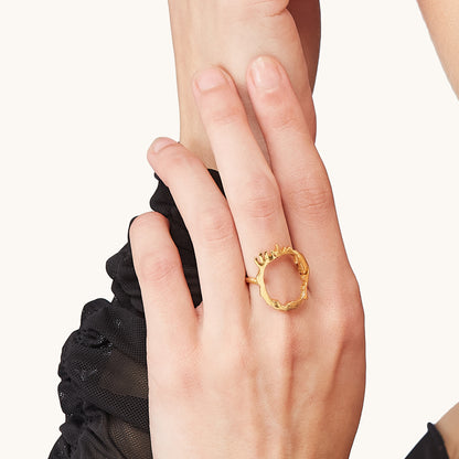 Close-up of a hand resting on an arm, wearing a statement yellow gold-plated ring with a hollow, textured molten-metal "drip" detailing along the top edge.