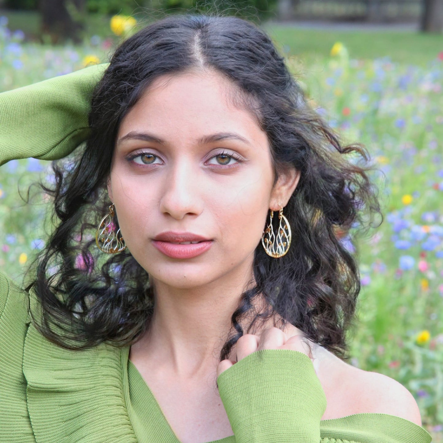 A model with dark, curly hair wearing intricate two-tone gold and silver circular drop earrings, posing in a field of colorful wildflowers while wearing a green sweater.