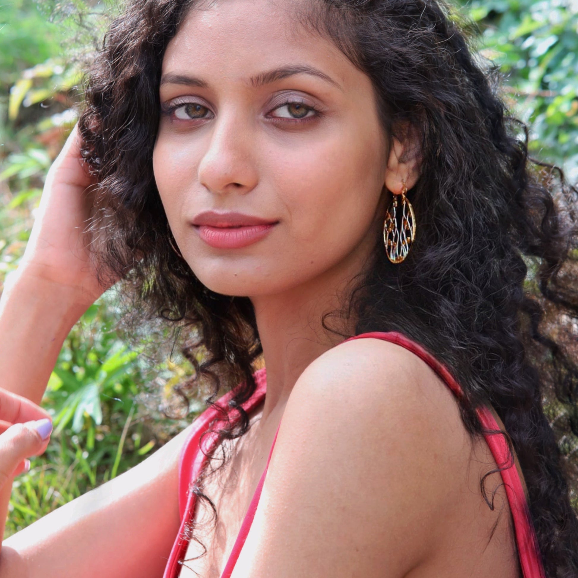 A portrait of a woman with dark curly hair looking directly at the camera, wearing sophisticated two-tone metallic openwork earrings.