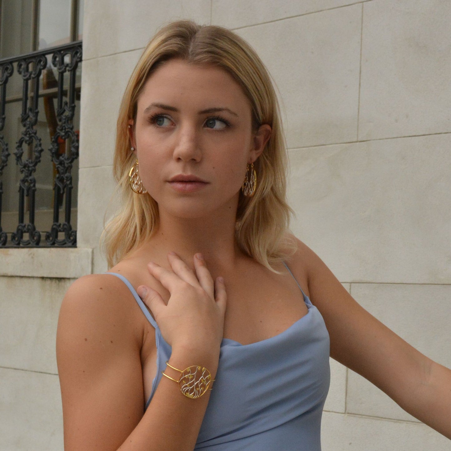 A blonde model wearing a light blue dress and a matching two-tone openwork cuff bracelet and circular drop earrings, looking off to the side against a neutral stone background.