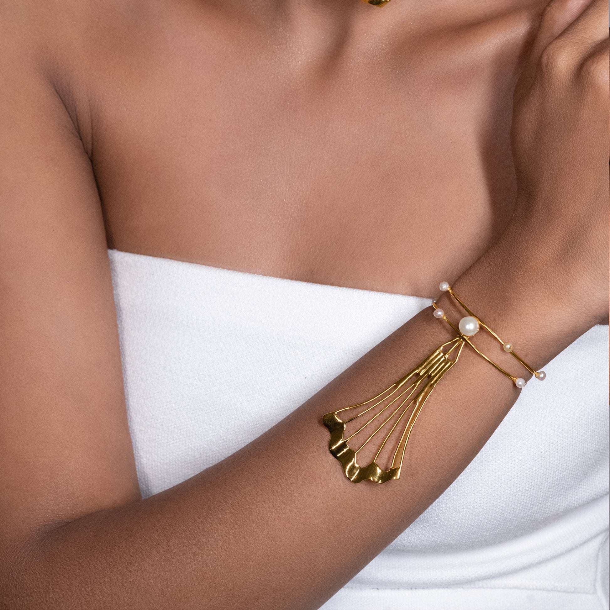 A close-up view of a model's arm wearing an elegant gold cuff bracelet adorned with delicate pearls and a sweeping, fan-like metal accent.
