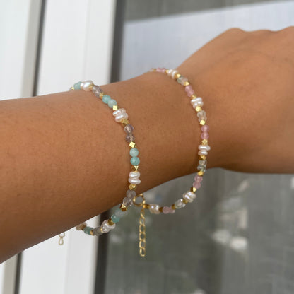 A close-up of a wrist wearing two delicate beaded bracelets, featuring a mix of pastel-colored stones, pearls, and gold-toned accents.