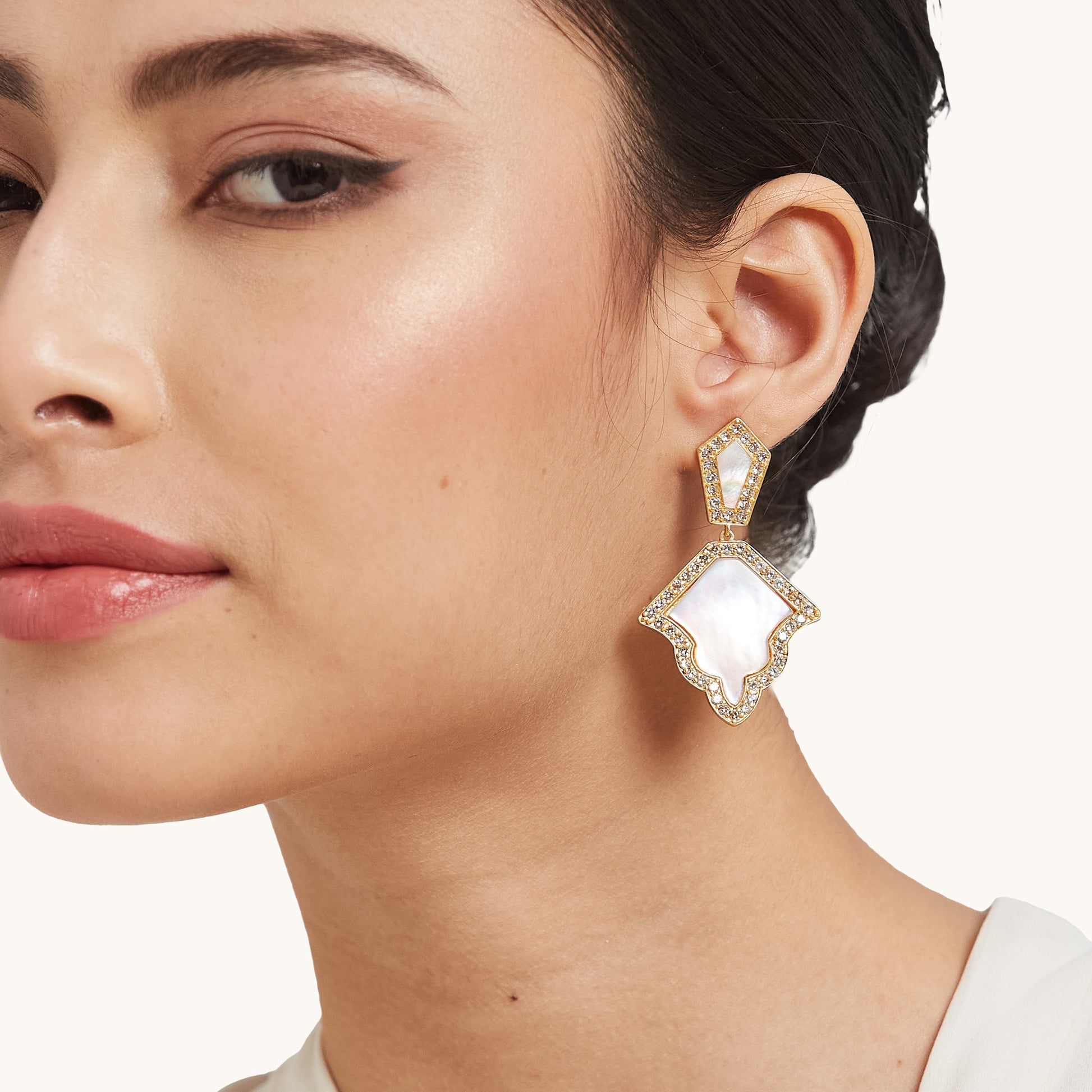 Close-up of a model wearing a luxurious geometric drop earring with an iridescent mother of pearl centerpiece with cubic zirconia accents  in gold setting.