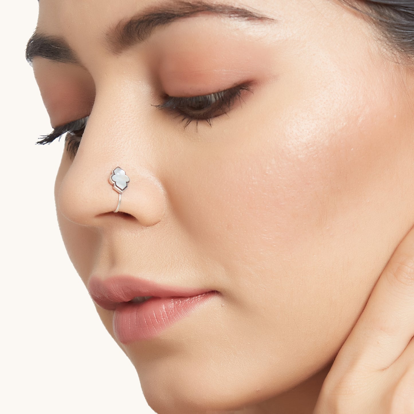 A close-up profile shot of a model wearing a small, decorative silver-toned clip-on nose ring with an elegant, white, four-lobed stone inlay.