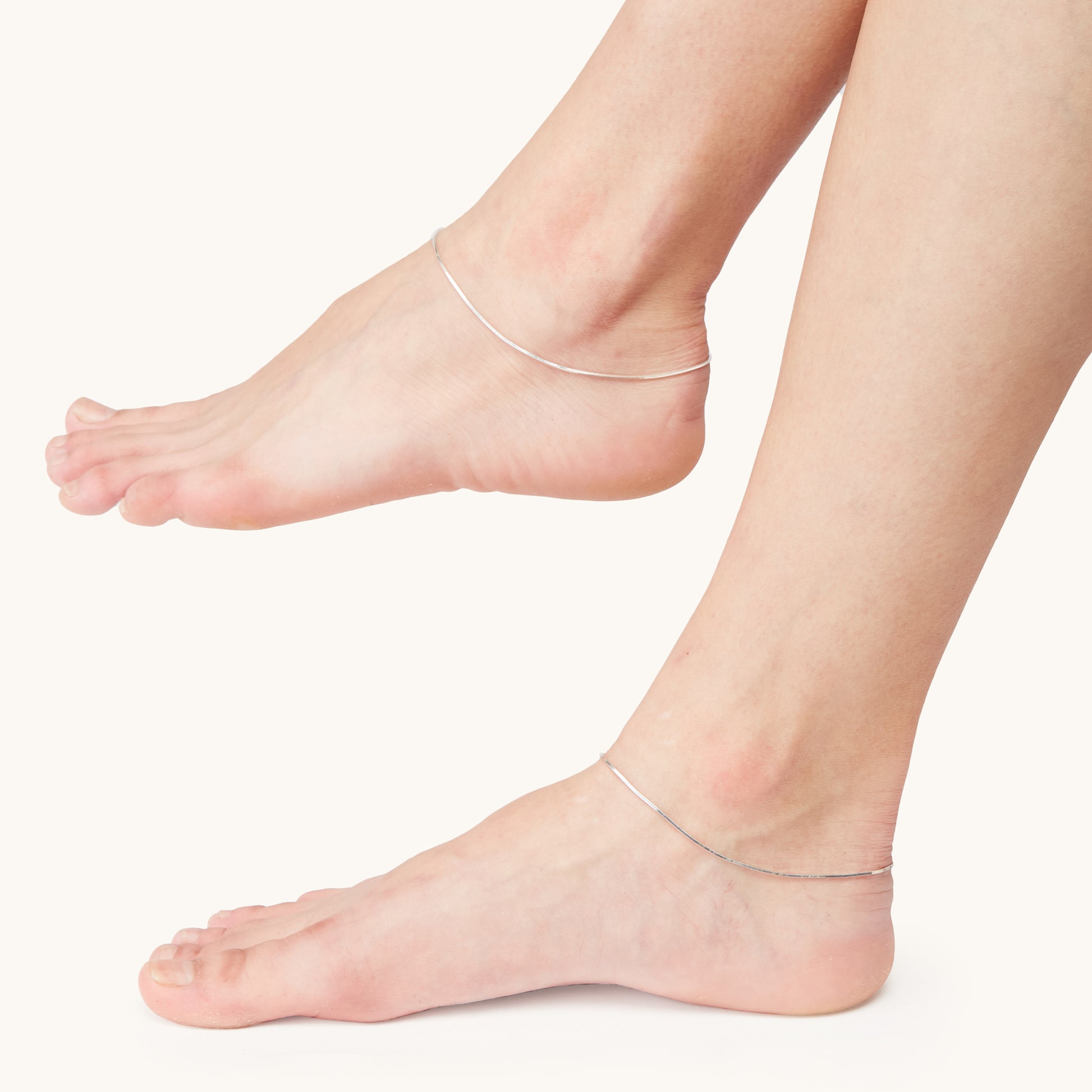 Detailed view of a minimalist silver chain anklet worn on a model's ankles against a bright white studio background.