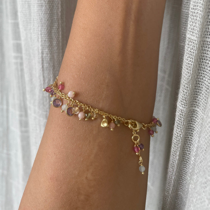 A close-up of a wrist wearing a dainty double-chain bracelet featuring dangling, multi-colored gemstone charms.