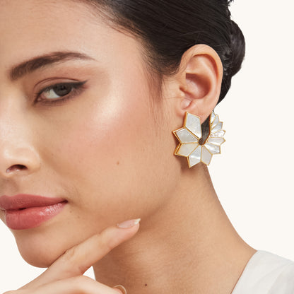 A close-up of a model wearing a large, fan-shaped gold earring with white shell inlays, tucked behind the ear with dark hair styled back.
