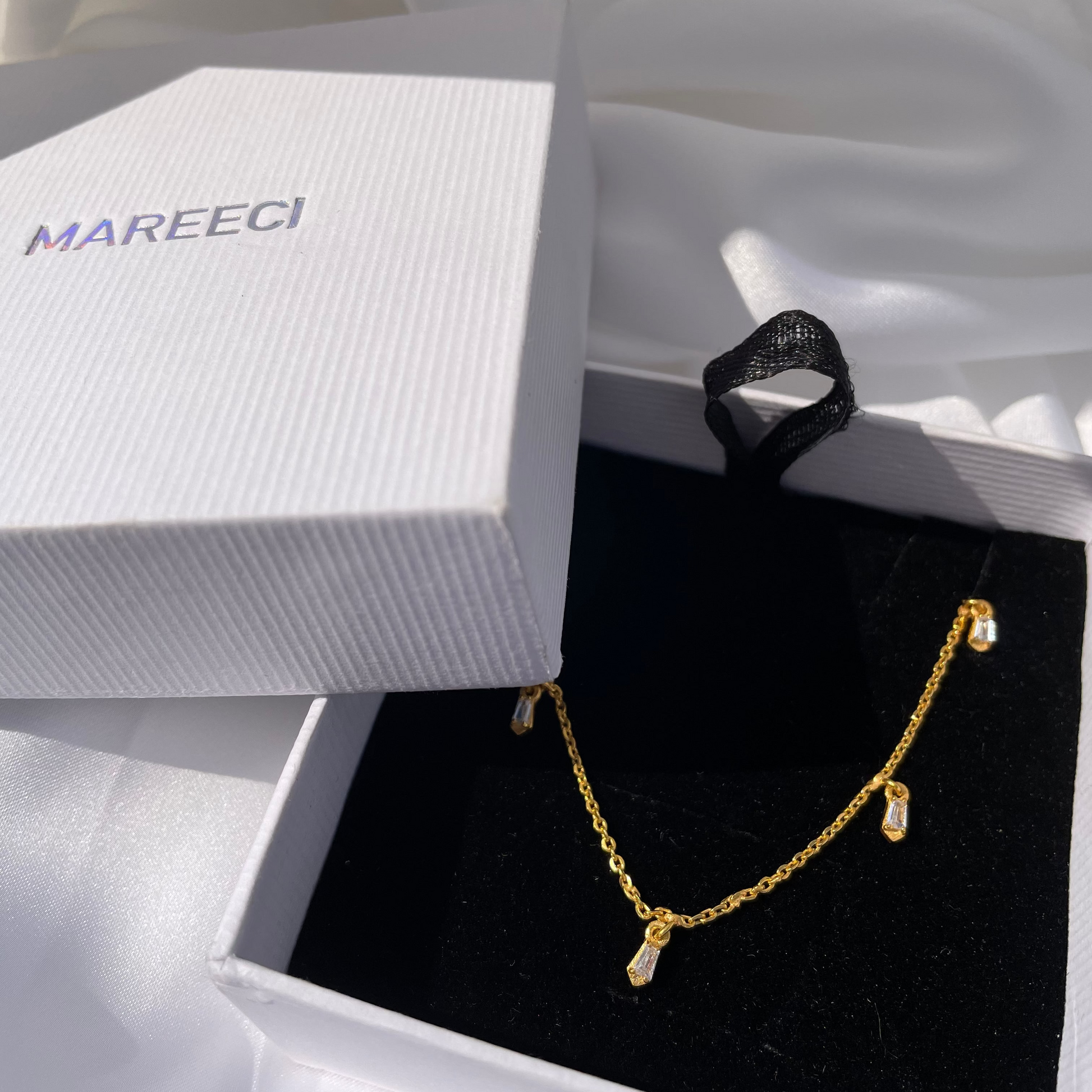 Elegant yellow gold floating charm necklace displayed on a black velvet insert inside a premium white Mareeci gift box, set against a soft white draped fabric background.
