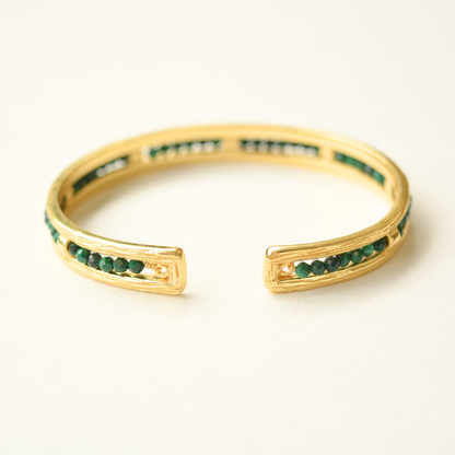 A close-up view of a gold-toned open cuff bracelet with emerald bead detailing set within a textured frame.