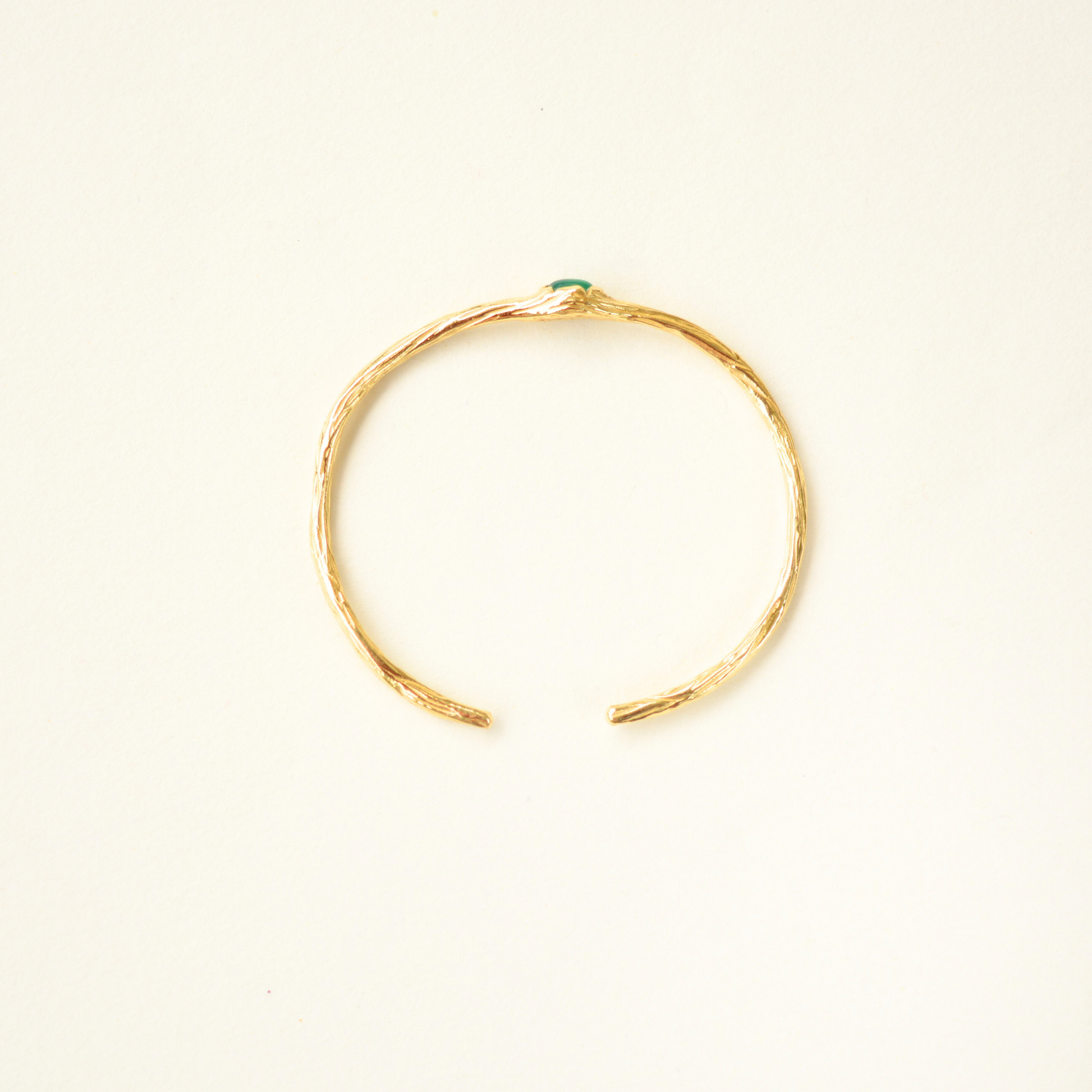 Top-view of a gold-plated cuff bracelet featuring a detailed, organic textured band and a center green stone.
