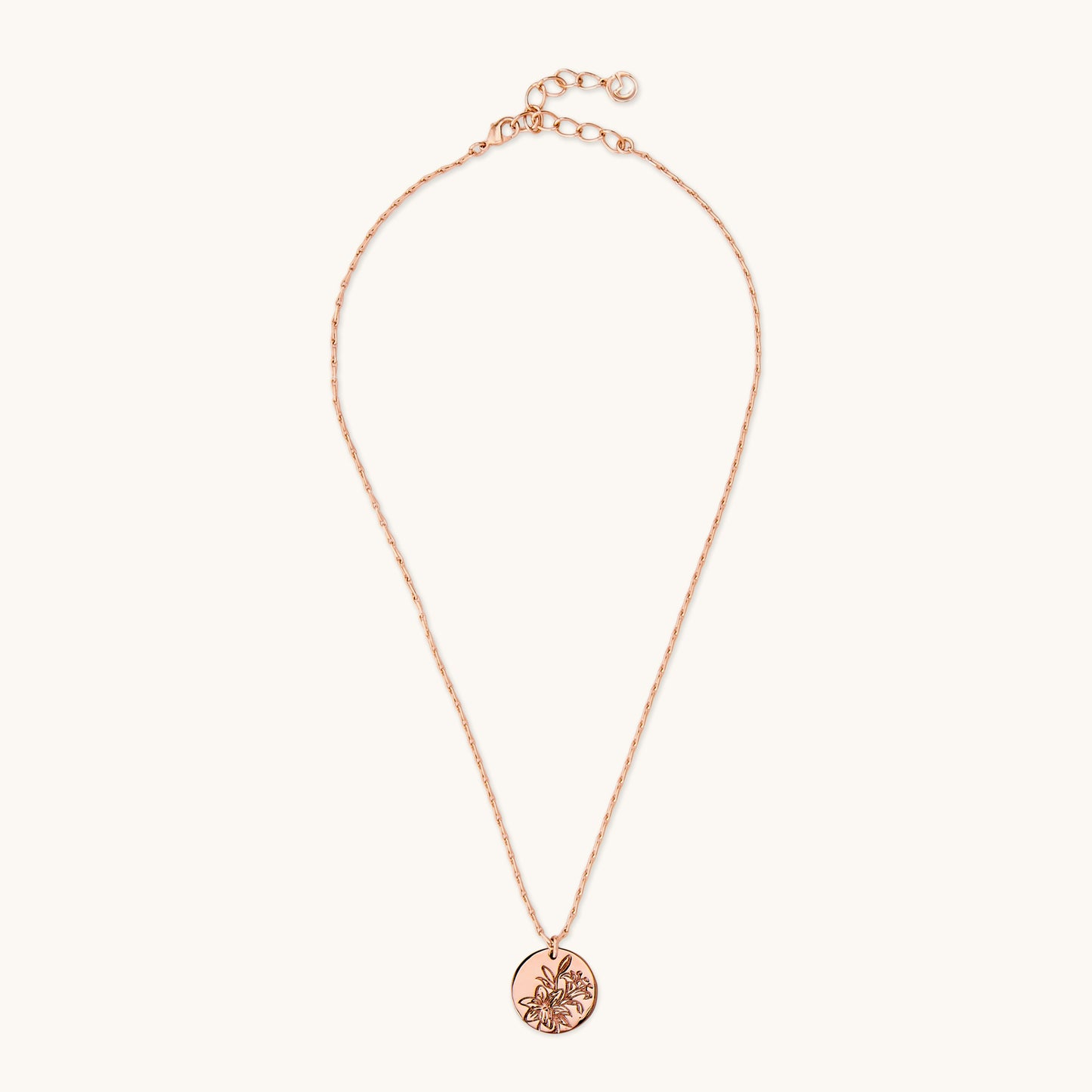 A polished circular pendant featuring an intricate floral lily engraving, suspended on a delicate brass rose gold chain against a plain white background. 