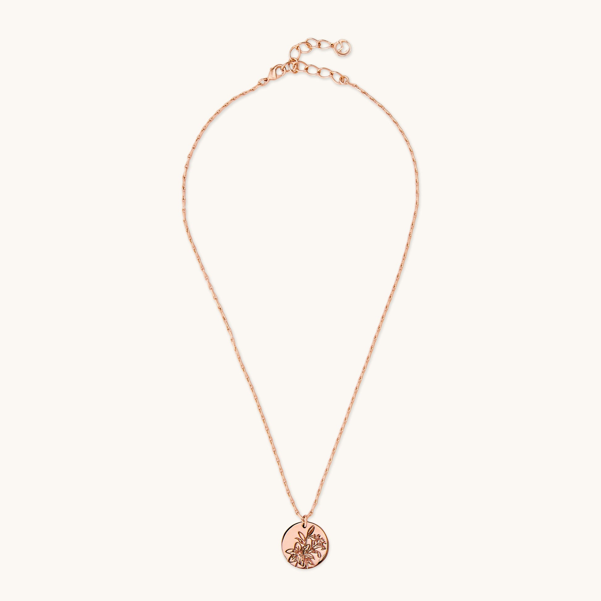 A polished circular pendant featuring an intricate floral lily engraving, suspended on a delicate brass rose gold chain against a plain white background. 