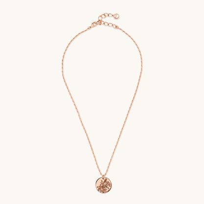 A polished circular pendant featuring an intricate floral lily engraving, suspended on a delicate brass rose gold chain against a plain white background. 