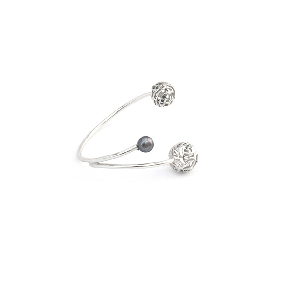 A silver, open-ended bracelet featuring two hollow, filigree-style spheres with  accents and one smooth pearl charm, isolated on a white background.