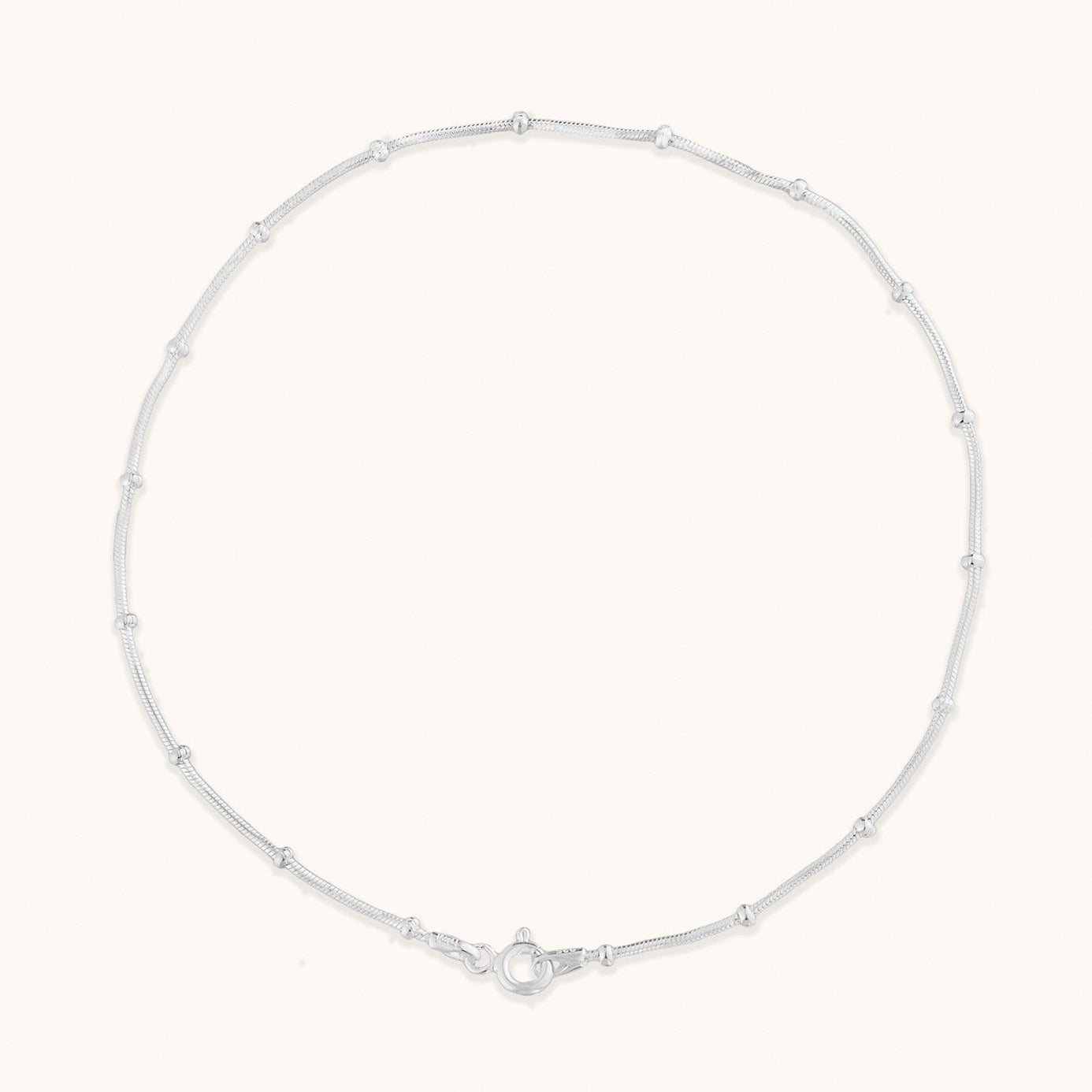A studio shot of a single minimalist silver-toned chain anklet with delicate bead accents, laid out in a circle to show its length and clasp detail.