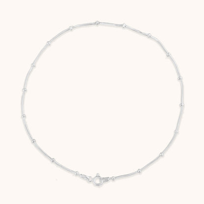 A studio shot of a single minimalist silver-toned chain anklet with delicate bead accents, laid out in a circle to show its length and clasp detail.