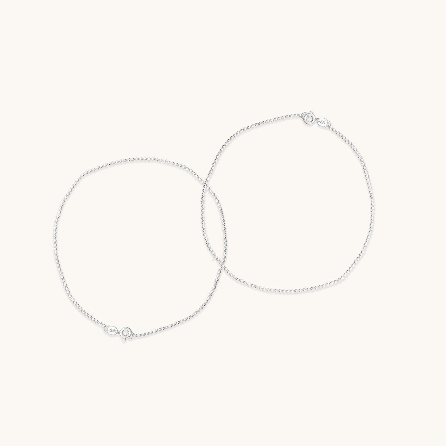 Two fine, shimmering silver chain anklet  positioned in overlapping circles to demonstrate their delicate texture and clasp hardware.